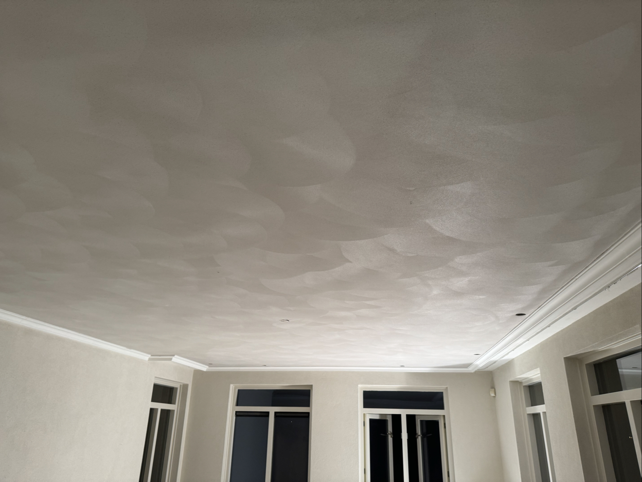 Strak gestuct wit plafond met sierlijst boven een kamer met hoge ramen